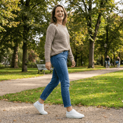 Lachende vrouw in beige trui en blauwe jeans loopt in een zonnig park met witte sneakers.